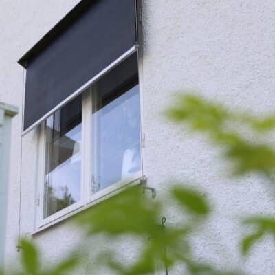 Svart halvt neddragen fönstermarkis på vit putsad fasad med gröna växter i förgrunden
