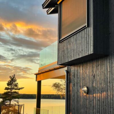 Svart fasad med neddragen vertikalmarkis vid solnedgång över sjö.